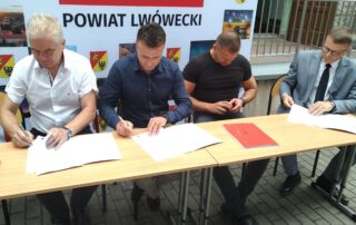 4 mężczyzn siedzących przy podłużnym stole na tle ścianki reklamowej Powiatu Lwóweckiego podpisuje dokumenty