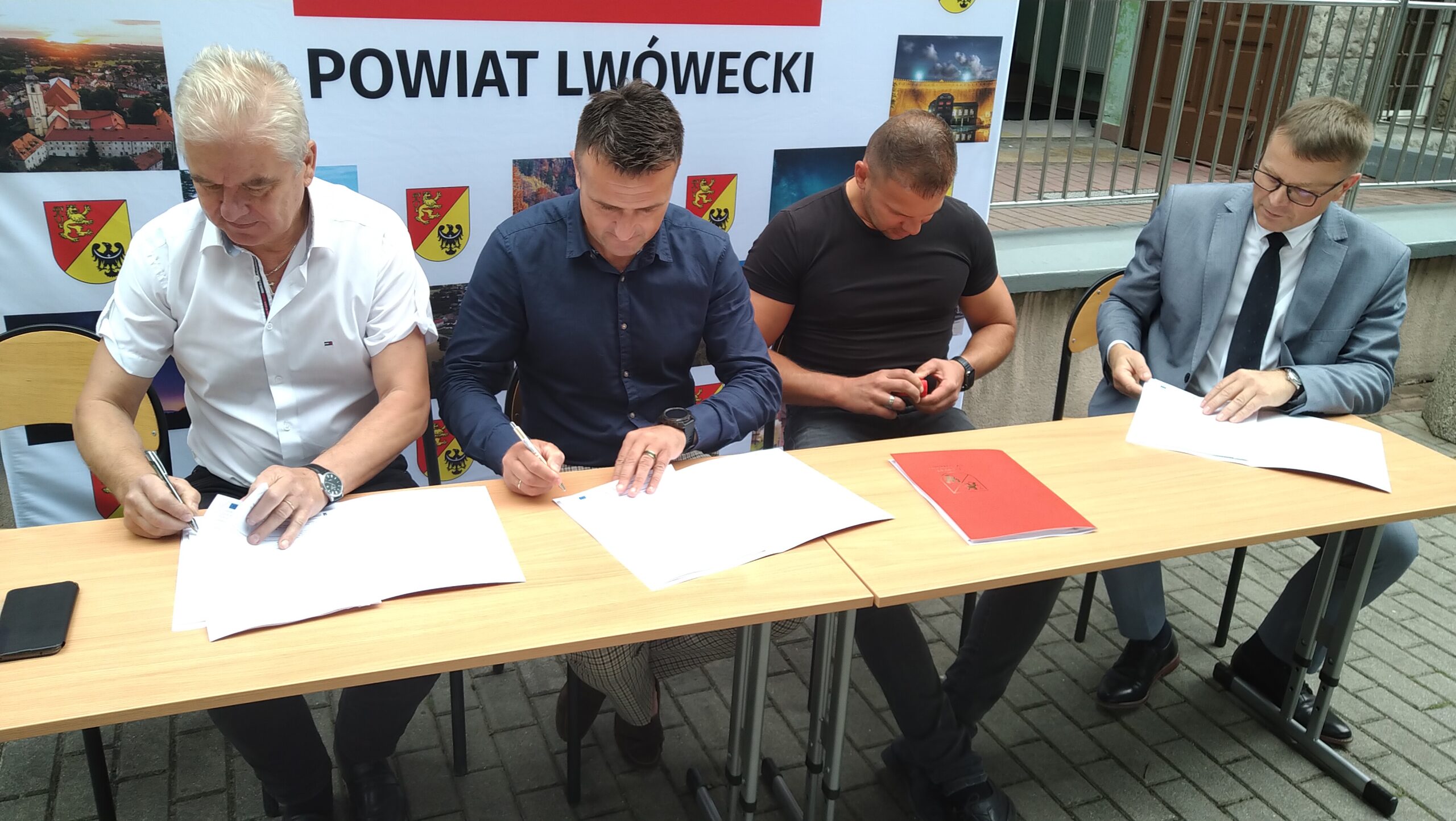 4 mężczyzn siedzących przy podłużnym stole na tle ścianki reklamowej Powiatu Lwóweckiego podpisuje dokumenty