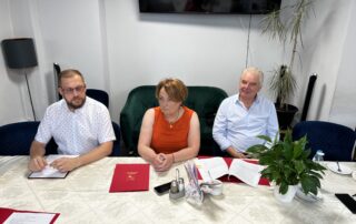 Na zdjęciu siedzący mężczyzna w okularach kobieta w pomarańczowej bluzce oraz mężczyzna w białej kosuzli przy stole na którym leżą dokumenty