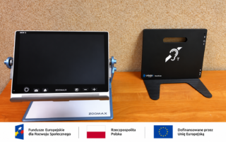 Na drewnianym blacie stoją dwa urządzenia zakupione w ramach projektu „Dostępny samorząd 2.0”: po lewej elektroniczny powiększalnik ZOOMAX Snow 12 z dużym ekranem, a po prawej czarna przenośna pętla indukcyjna Univox SmartLoop oznaczona symbolem dostępności dla osób słabosłyszących. Sprzęt przeznaczony jest dla Powiatowego Zespołu ds. Orzekania o Niepełnosprawności.