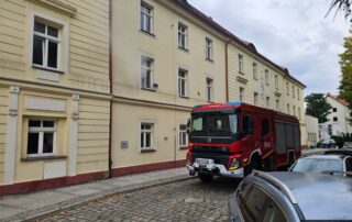 Czerwony samochód Państwowej Straży Pożarnej zaparkowany przed budynkiem Starostwa Powiatowego w Lwówku Śląskim podczas ćwiczeń ewakuacyjnych z zakresu ochrony ludności i obrony cywilnej.